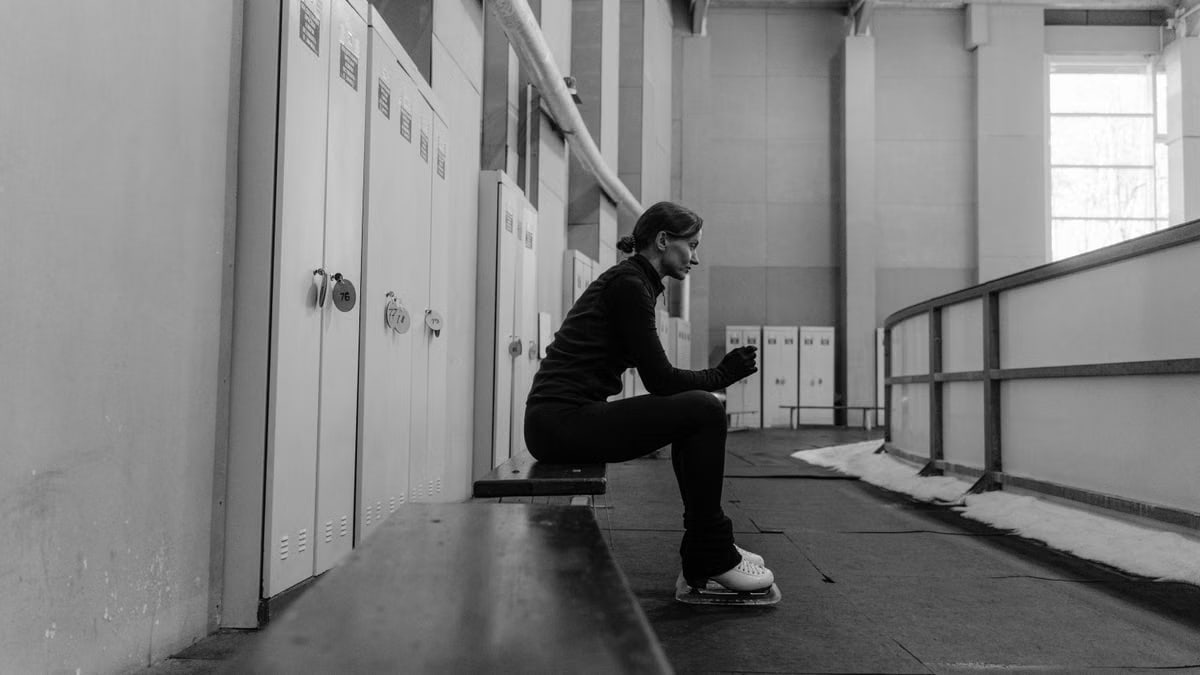 A female figure skater resting on a bench in an indoor rink locker area, in a black and white setting.